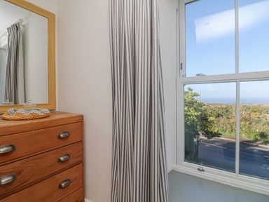 A wooden chest of drawers with a mirror and striped curtains next to a window showing a road and greenery at Bal View in St Just