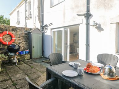 An outdoor patio with a table set with mugs croissants and a teapot near a sliding glass door and a barbecue grill at Bal View in St Just