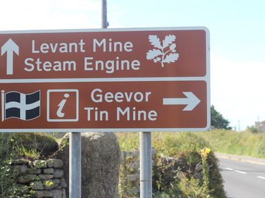 A brown road sign indicating directions to Levant Mine Steam Engine and Geevor Tin Mine by a roadside with vegetation and stone wall