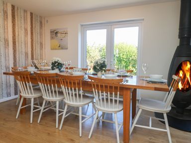 A dining room with a long wooden table set for eight with white chairs a black fireplace and glass doors at Penhaven in Crackington Haven