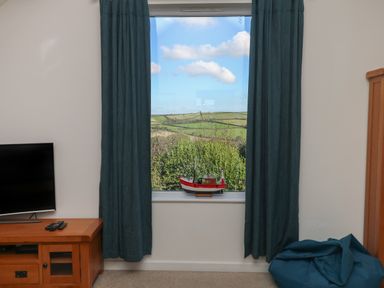 A window with blue curtains a model boat on the windowsill a television on a wooden stand and a beanbag chair in a room at Penhaven in Crackington Haven