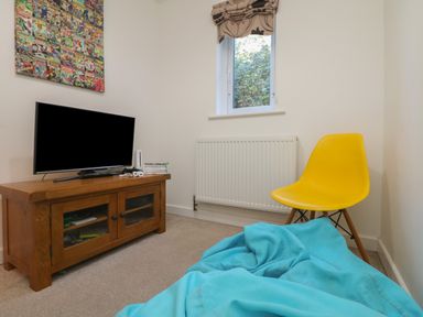 A small room with a wooden TV stand holding a flat screen TV and video games a yellow chair a window with a patterned blind and a blue bean bag on the floor at Penhaven in Crackington Haven