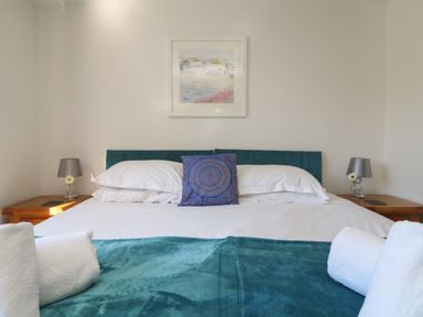 A bedroom with a double bed with white pillows and a teal blanket two bedside tables with lamps and flowers and a framed picture on the wall at Penhaven in Crackington Haven