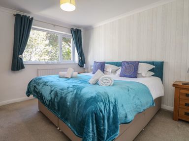 A bedroom with a large bed with blue blanket and pillows beside a window with blue curtains and a wooden nightstand at Penhaven in Crackington Haven