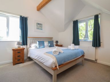 A bedroom with a bed with blue pillows and blanket two windows with blue curtains two wooden bedside tables with lamps and a small boat decoration on the window at Penhaven in Crackington Haven