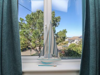 A window with green curtains and a small sailboat model on the windowsill overlooking trees and houses at Penvahen in Crackington Haven