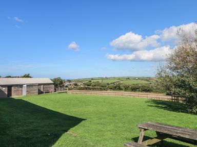 An outdoor space with a wooden picnic table and a building beside a grassy field and rural hills at Penhaven in Crackington Haven