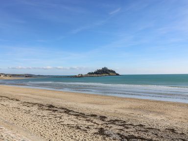 A sandy beach with seaweed patches and calm waves with an island and buildings in the distance at Mackerel Sky in Marazion