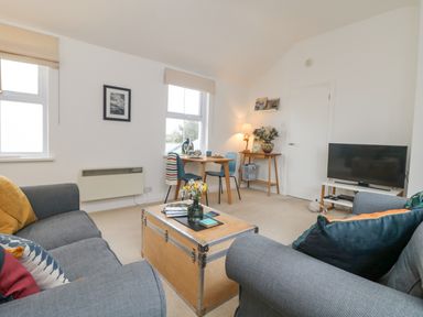 A living room with two gray sofas a wooden coffee table a small dining table with two chairs a TV on a stand and a side table with a lamp and flowers at Mackerel Sky in Marazion
