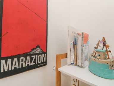 A corner of a room with a red Marazion poster on the wall a white table with books and a decorative boat model on it at Mackerel Sky in Marazion
