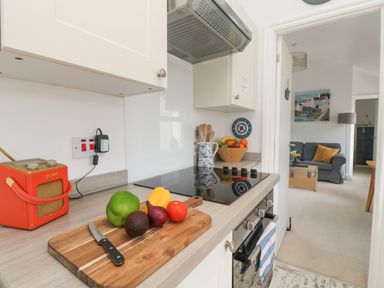 A kitchen countertop with a chopping board holding a knife and assorted vegetables next to an electric stove and a red radio with a glimpse of a sitting room with a sofa and table at Mackerel Sky Marazion