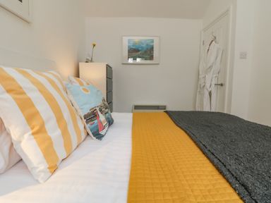 A bedroom with a bed with yellow and white striped pillows a blue pillow a yellow blanket a gray throw a chest of drawers a white door with robes hanging and a framed picture on the wall at Mackerel Sky in Marazion