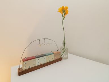 A decorative model of houses with birds hanging above next to a tall yellow flower in a glass vase on a white surface at Mackerel Sky in Marazion