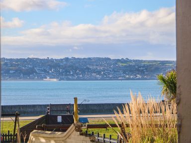 A seaside view with a playground and bench in the foreground and houses on a hillside across the water at Mackerel Sky in Marazion