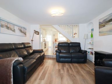 A living room with black leather sofas, wooden floor, floor lamps, paintings, and an open view to a kitchen and dining area at Alma House 2 in Perranporth