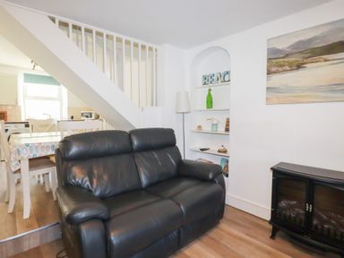 A living room with a black leather sofa a dining table under stairs a standing lamp and shelves with decorations at Alma House 2 in Perranporth