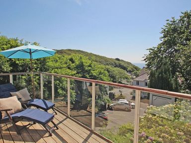A balcony with two lounge chairs blue cushions and a blue umbrella overlooking trees and houses at Porthcurno Bay View in Porthcurno