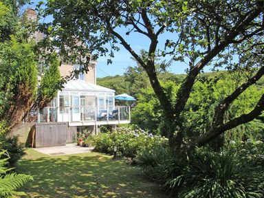 A garden with trees and bushes near a house with a glass conservatory and patio furniture at Porthcurno Bay View in Porthcurno