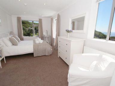 A bedroom with a bed a dresser a white armchair and windows showing a view of greenery and water at Porthcurno Bay View in Porthcurno