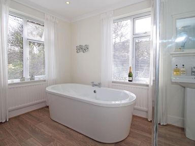 A bathroom with a freestanding bathtub between two windows with blinds and sheer curtains at Porthcurno Bay View in Porthcurno