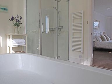 A bathroom with a white bathtub, glass shower enclosure, towel rack, and a small shelf with towels and a vase with flowers at Porthcurno Bay View in Porthcurno