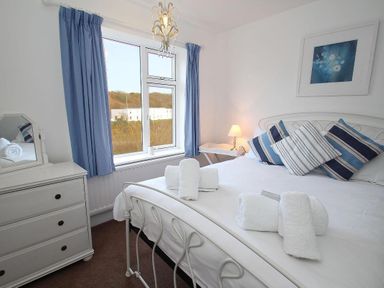 A bedroom with a white metal bed frame blue curtains a white dresser and a bedside table with a lamp at Porthcurno Bay View in Porthcurno