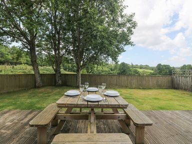 A wooden picnic table set with plates and glasses on a deck overlooking a fenced grassy garden with trees at Willow in Falmouth