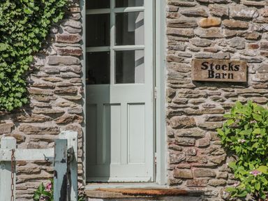 A stone wall with a pale green wooden door partly open and a sign reading Stocks Barn with green plants nearby at Stocks Barn in Looe