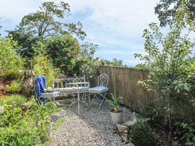 A garden seating area with white metal chairs and table on a gravel path surrounded by plants at Stocks Barn in Looe