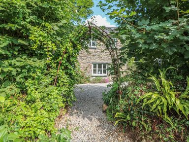 A gravel pathway leading to a stone house with two windows surrounded by green plants and an arch covered in climbing vines at Stocks Barn in Looe