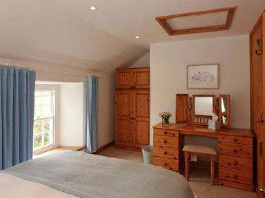 A bedroom with a bed near a window with blue curtains a wooden wardrobe and a wooden dressing table with a mirror and stool at Halvosso in Falmouth