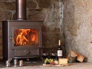 A wood burning stove with fire inside near a stone wall with wine, two glasses, grapes, figs, cheese, bread, and logs at Halvosso in Falmouth