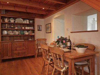 A dining area with a wooden table set with plates wine and a bowl of salad wooden chairs a wooden cabinet with dishes and cups on shelves and two framed pictures on the wall at Halvosso in Falmouth