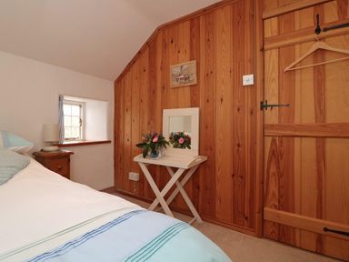 A bedroom with wooden paneled walls a bed a bedside table lamp a small window and a folding table with a mirror and flowers at Halvosso in Falmouth