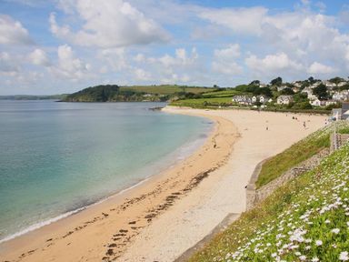 A sandy beach beside calm water with houses and green fields in the background at Halvosso in Falmouth
