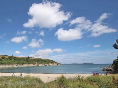 A sandy beach with grass in the foreground and two people sitting near the water with hills and blue sky at Halvosso in Falmouth