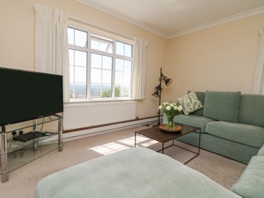 A living room with a green sofa, a black coffee table with flowers, a flat screen tv on a glass stand and a large window at Charlesworth in Braunton