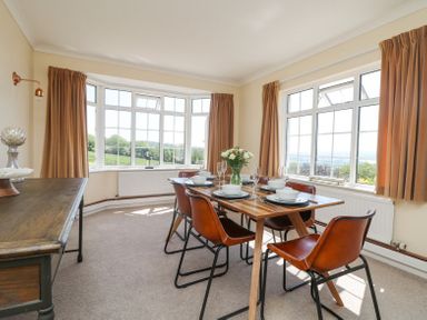 A dining room with a wooden table and six leather chairs next to large windows with beige curtains at Charlesworth in Braunton