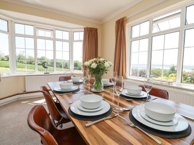 A dining room with a wooden table set with white dishes chairs and large windows showing greenery at Charlesworth in Braunton