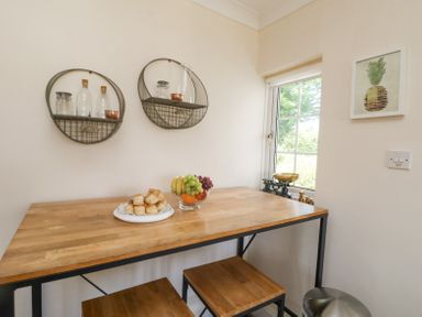 A wooden table with pastries and a bowl of fruit two wooden stools a window with a scale on the sill and round wall shelves with glass jars and bottles a framed pineapple print on the wall at Charlesworth in Braunton