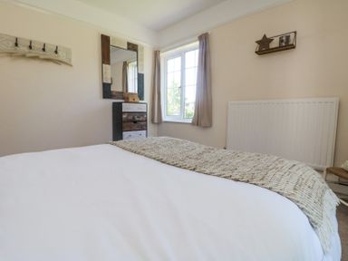 A bedroom with a bed white blankets a window with curtains a radiator a chest of drawers with a mirror and wall decorations at Charlesworth in Braunton
