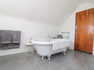 A bathroom with a freestanding clawfoot bathtub a white towel on the tub a small round side table with a bottle two gray towels on a wall rack and a wooden door at Charlesworth in Braunton