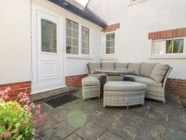 An outdoor patio with a white door and windows a gray wicker corner sofa set with a glass table at Charlesworth in Braunton