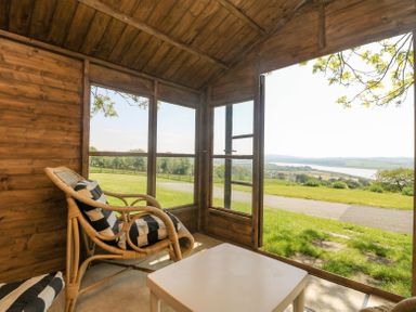 A wooden porch with a wicker chair with black and white cushions and a white table overlooking a grassy landscape and a river at Charlesworth in Braunton
