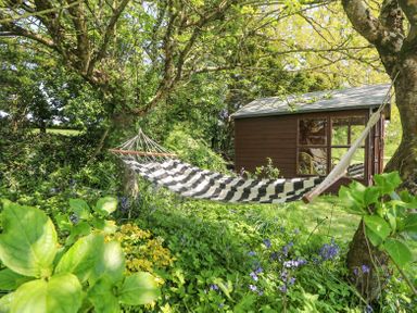 A black and white striped hammock tied between two trees in a garden with plants and flowers next to a wooden shed at Charlesworth in Braunton