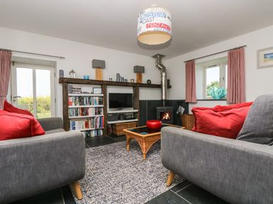 A living room with two gray sofas with red cushions a wood burning stove a bookshelf and a glass coffee table at Sundowner in Padstow