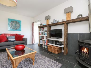 A living room with a gray sofa red cushions a wicker coffee table with a red bowl a wood burning stove and a wooden shelf with a television and books at Sundowner in Padstow