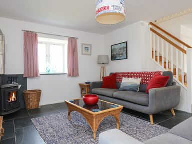 A living room with a grey sofa red cushions a wicker coffee table with a red bowl a wood burning stove and stairs in the background at Sundowner in Padstow