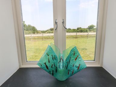A decorative glass vase with fish designs on a windowsill overlooking a grassy yard and fence at Sundowner in Padstow