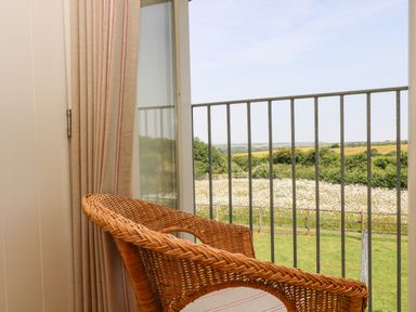 A wicker chair next to a glass door with beige curtains overlooking a garden and field at Sundowner in Padstow